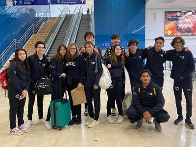 A group of students in dark hoodies, with suitcases, poses by an escalator at an airport.