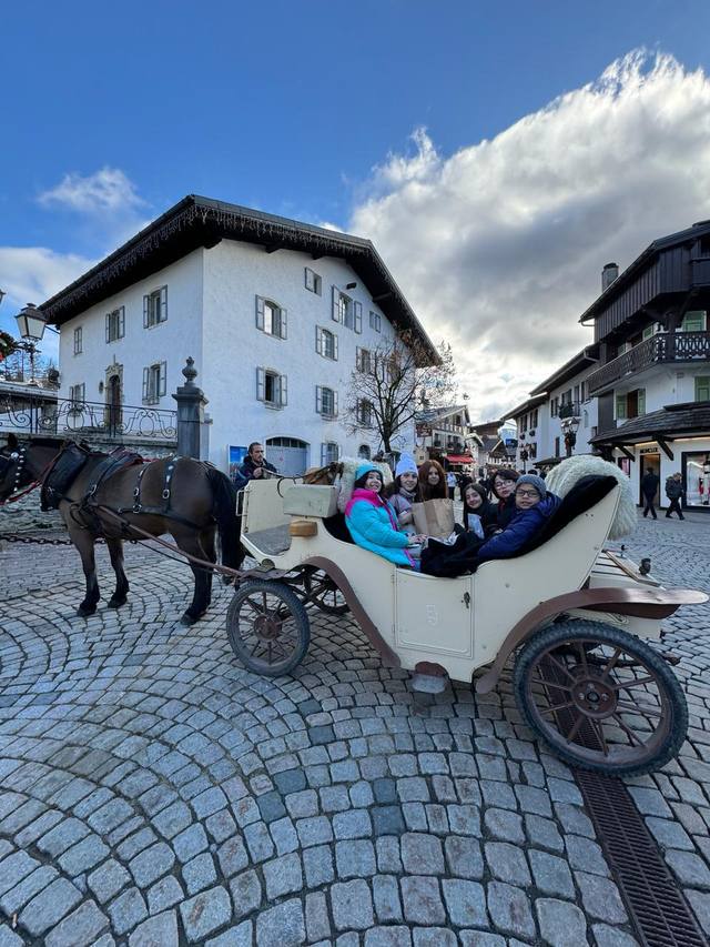 Smiling students ride in a horse-drawn carriage through a cobblestone square lined with Alpine houses.