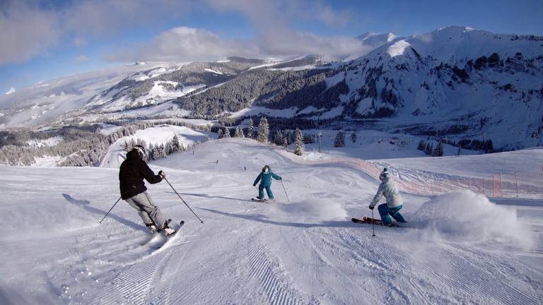 Three skiers head down a freshly groomed slope, with a snowy valley and tall mountains behind them.