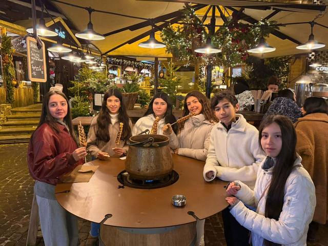 Six students with skewers gather around a fondue pot at a Christmas market lit up at night.