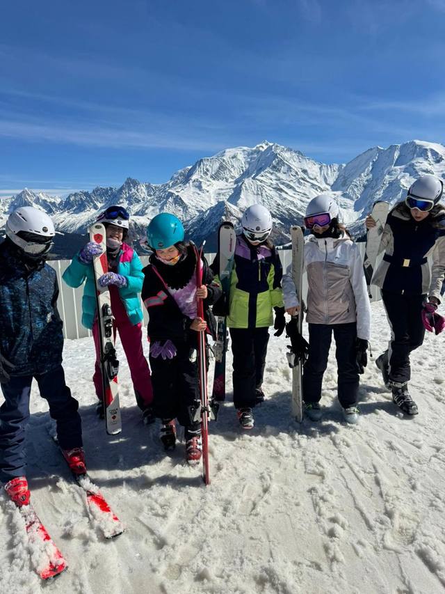 Six students in helmets stand in the snow holding their skis, with snow-covered mountains behind them.