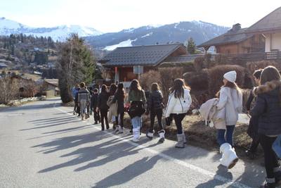 A group of students, seen from behind, walks along a mountain road between chalets toward snowy peaks.
