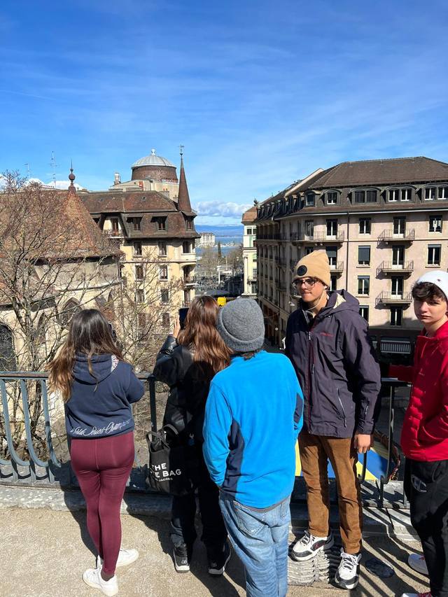 Students stand at a city lookout with a railing, between old buildings, with a lake in the distance.