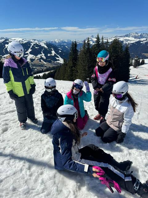 Six students in helmets sit and rest in the snow at the top of a slope, with a valley and mountains behind them.