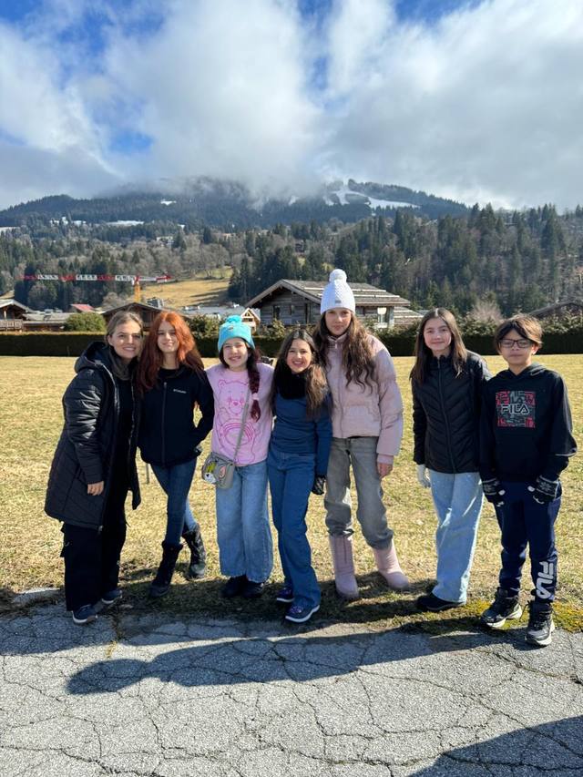 Seven students pose together in a meadow, with mountains, forest, and clouds behind them.