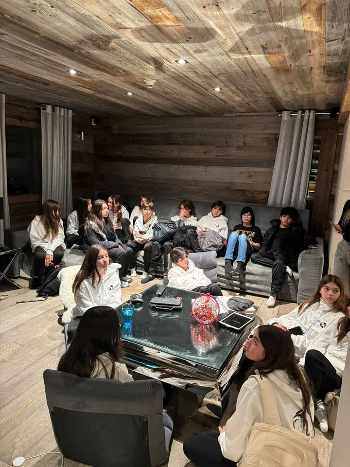 About twenty students in white MAcC hoodies gather at night in a room with a wooden ceiling and walls.