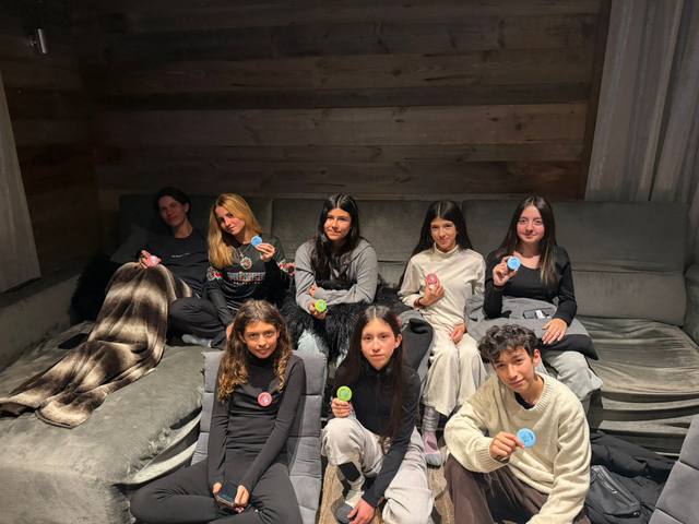 Nine students in pajamas and comfy clothes sit on a couch in a wood-paneled room, showing off colorful pins.