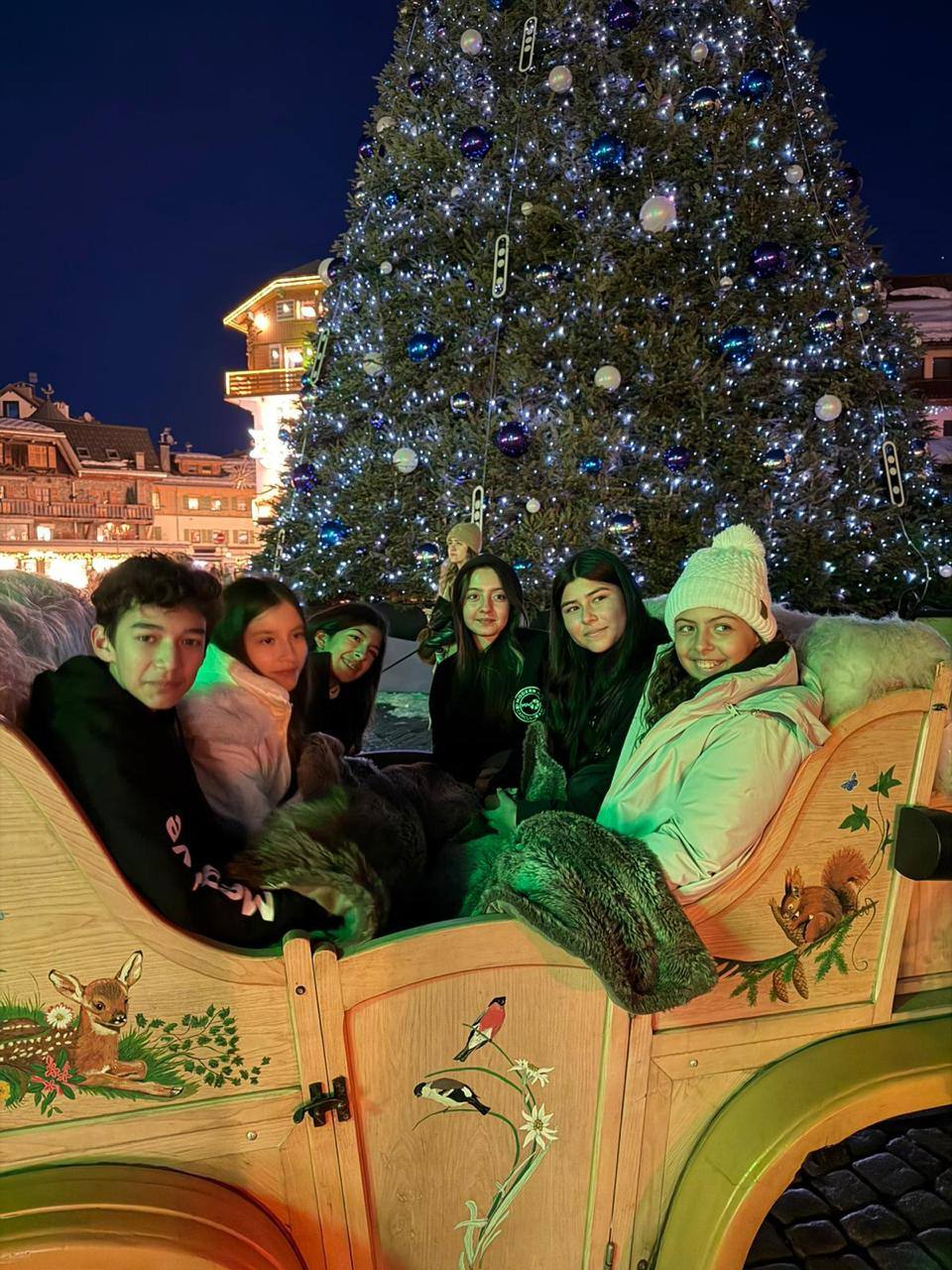 Six students sit in a painted wooden sleigh at night, in front of a big lit-up Christmas tree.